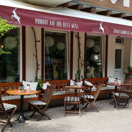 Restaurant mit Tischen und Stühlen im Freien, umgeben von Pflanzen und einer einladenden Atmosphäre.