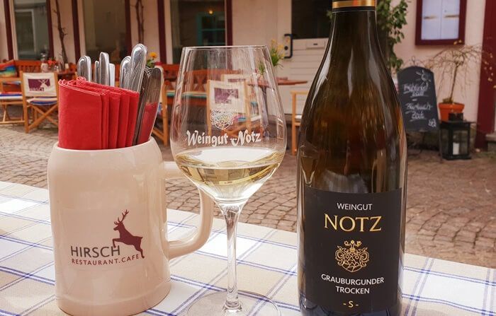 Eine Flasche Grauburgunder vom Weingut Notz, ein gefülltes Weinglas und ein Gefäß mit roten Servietten und Besteck.