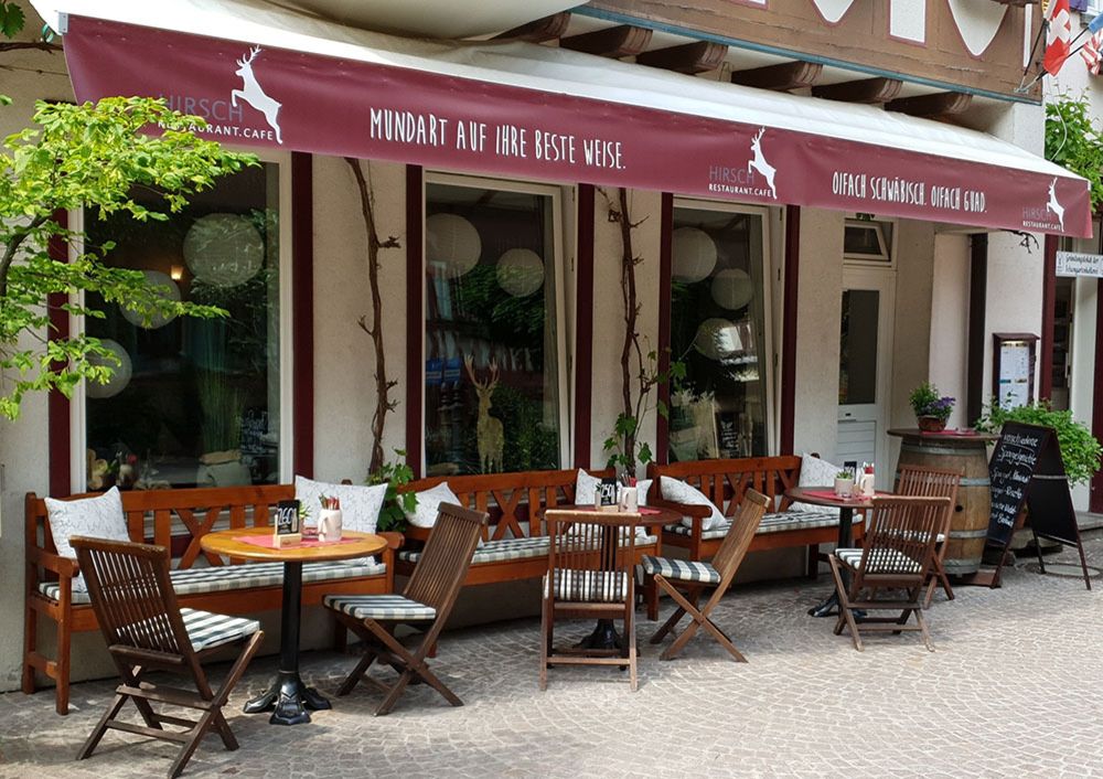 Außenbereich des Restaurants Hirsch | Rotes und weißes Markisen, die über einem Eingang gespannt sind, bieten Schatten und Schutz vor Sonne und Regen.